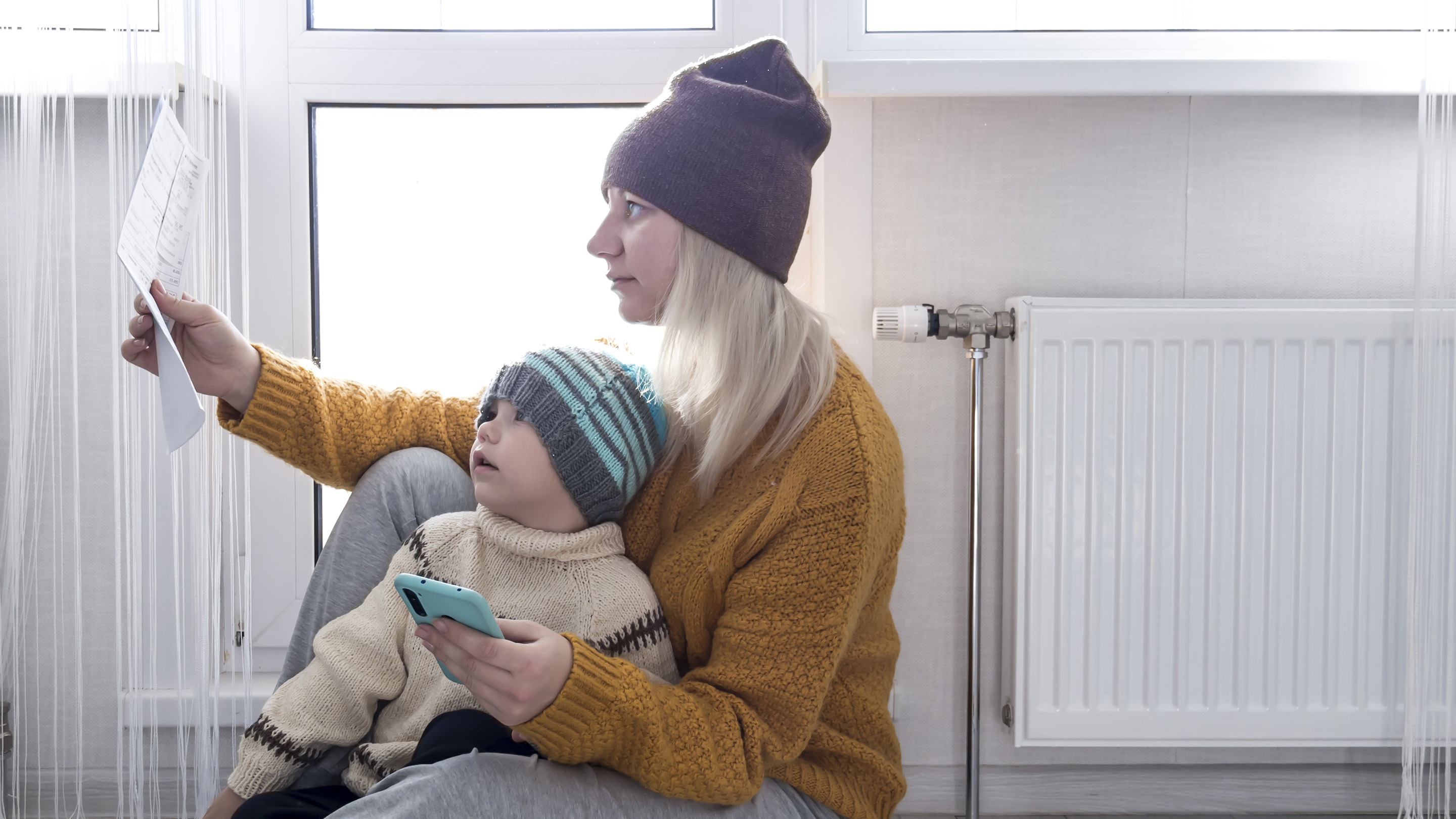 .Una madre con un niño pequeño con un suéter amarillo y sombreros está contando dinero y pensando en cómo pagar facturas e impuestos, cerca de un calentador con un termostato.